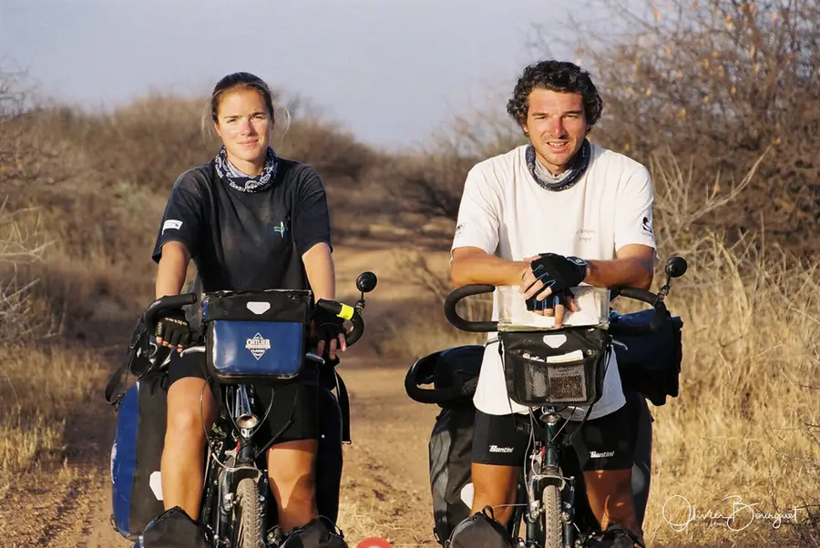 Deux cyclistes souriants appuyés sur leurs vélos chargés, sur une piste de brousse